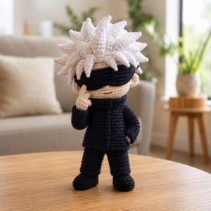May include: A handcrafted crochet doll featuring a character with spiky white hair, a black blindfold covering the eyes, and a light beige complexion. The figure wears a dark navy blue high-collared jacket and matching trousers with black shoes.