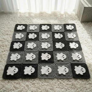 May include: A square crochet blanket featuring a grid of alternating dark grey and black squares. Each square contains a white ghost applique with two small black eyes and a smiling mouth, creating a repeating pattern across the entire textile.