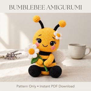 Op de afbeelding: Een gehaakt hommel amigurumi speeltje met gele en zwarte strepen, zwarte voelsprieten met gele uiteinden, roze wangen en grote zwarte ogen. Het houdt een wit madeliefje met een geel hartje vast en heeft een witte bloem. Tekst BUMBLEBEE AMIGURUMI.