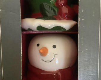 May include: A ceramic snowman head figurine with a white face, orange carrot nose, and black eyes, wearing a dark red scarf. Above it sits a green ceramic piece featuring a red cardinal bird perched on a white snow-covered base.