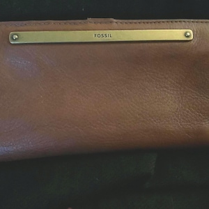 May include: Brown leather wallet with a rectangular gold-colored metal bar across the top. The word "FOSSIL" is embossed on the metal bar. The wallet is closed and has a textured surface.