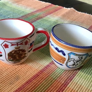 May include: Two white ceramic mugs with colorful hand-painted designs. One mug has a red rim and handle, with a crest featuring a brown hedgehog. The other has a blue rim and handle, with a crest featuring a horse.