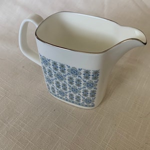 May include: A white ceramic creamer with a blue and green floral pattern. The creamer has a square body and a silver rim.