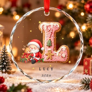 May include: A circular clear glass Christmas ornament featuring a large letter L shaped like a pink frosted cookie decorated with a candy cane and gingerbread man. Beside it is a cartoon Santa Claus and a reindeer. The name LUCY and 2026 are printed.