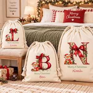 Pode incluir: Três sacos de presente de lona cor de creme com cordão, com letras grandes vermelhas L, B e N decoradas com azevinho e laços vermelhos. Cada saco apresenta uma pequena ilustração de uma raposa com gorro de Natal, o texto Merry Christmas e um nome.