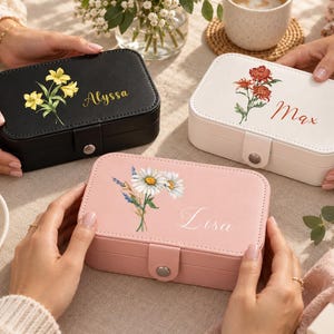 May include: Three rectangular jewelry boxes in black, white, and pink, each featuring a snap closure and a floral illustration. The black box is labeled with the name Alyssa, the white box with Max, and the pink box with the name Lisa in script.