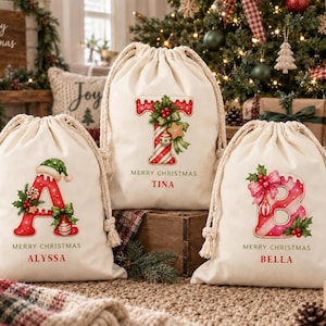Pode incluir: Três sacos de lona com cordão em tom off-white com gráficos festivos de Natal. Cada saco exibe uma letra grande, o texto MERRY CHRISTMAS e um nome personalizado: ALYSSA, TINA ou BELLA. As letras são decoradas com azevinho, laços e ornamentos.