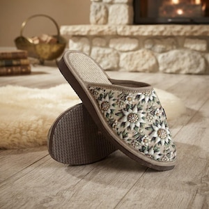 Op de afbeelding: Een paar instapslippers met een beige bovenkant voorzien van een wit en groen bloemenpatroon, een lichtgekleurde textuur binnenzool en een donkerbruine rubberen zool met een geribbeld profiel voor grip op binnenvloeren.