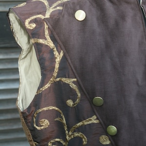 May include: A brown vest with gold embroidery. The vest has a high collar and is made of a textured fabric. The vest has several gold buttons.