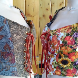 Peut inclure: Deux corsets en tissu brun et gris avec des lacets en cuir rouge et des œillets dorés. Un corset a un motif floral avec des citrouilles et des tournesols. L'autre corset a un motif plus abstrait avec des feuilles et des volutes.