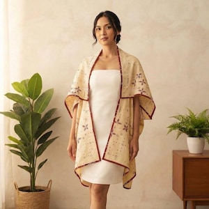 Könnte beinhalten: Ein cremefarbenes ärmelloses Kleid kombiniert mit einem vorne offenen Umhang, der ein zartes braunes Blumenmuster und einen dunkelbraunen Saum an den Kanten aufweist. Der Umhang fällt über die Schultern, während das Kleid schlicht geschnitten ist.