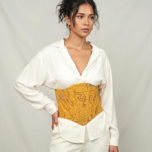 Può includere: Un corsetto giallo senape con un motivo floreale e geometrico marrone scuro indossato sopra una camicia bianca a maniche lunghe e pantaloni bianchi. Il corsetto presenta un orlo a punta strutturato e cuciture verticali.