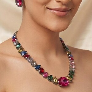 May include: A necklace featuring a series of multi-coloured, faceted, teardrop-shaped stones in shades of pink, blue, green, and yellow, set in a dark metal frame, paired with matching oval-shaped stud earrings with a central pink stone.