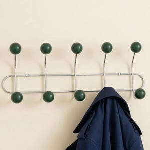 Può includere: Un appendiabiti da parete con una struttura orizzontale in metallo cromato e cinque supporti verticali. Ogni supporto ha una sfera verde scuro sia in alto che in basso, per un totale di dieci sfere. Un cappotto blu scuro pende da un gancio.