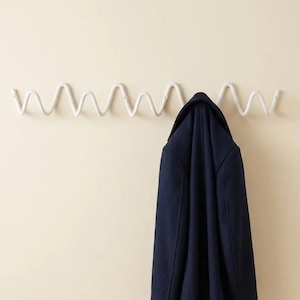 Pode incluir: Um cabide de parede branco com um padrão contínuo e repetitivo de ziguezague ou ondas. O cabide é construído a partir de uma única peça de metal e é mostrado com um casaco azul-marinho escuro pendurado num dos ganchos à direita.