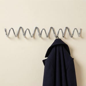 Op de afbeelding: Een wandkapstok met een doorlopend frame van gepolijst chroommetaal in een herhalend zigzag- of golfpatroon. Een donkerblauwe jas hangt aan een van de metalen haken, wat het functionele ontwerp van de wandbevestiging goed laat zien.