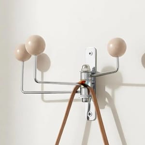 Peut inclure: Un porte-manteau mural composé d'une base verticale en métal chromé avec trois bras rotatifs. Chaque bras se termine par une sphère en bois beige clair lisse. Une lanière en cuir marron est suspendue au pivot central de la structure métallique.