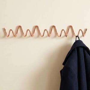 Op de afbeelding: Een roségouden wandkapstok met een doorlopend, herhalend zigzag- of golfachtig metalen draadontwerp. Een donkerblauwe jas hangt aan de rechterkant van de kapstok, die met zichtbare schroeven aan de muur is bevestigd.