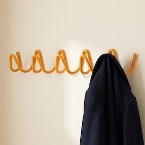 Peut inclure: Un porte-manteau mural jaune vif doté d'un design en fil métallique ondulé continu avec plusieurs crochets arrondis. Un manteau bleu marine foncé est suspendu à l'un des crochets, masquant partiellement le côté droit de la structure métallique.
