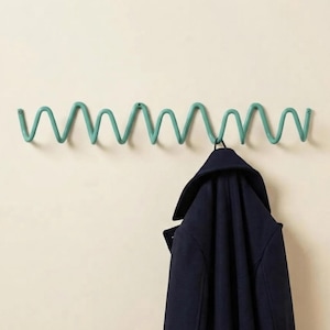 Op de afbeelding: Een aan de muur gemonteerde kapstok in blauwgroen met een doorlopend golvend, zigzag metalen ontwerp met meerdere haken voor het ophangen van items. Een donkerblauwe jas hangt aan een van de haken aan de rechterkant van de metalen structuur.