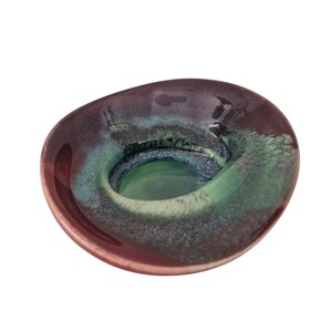 May include: A small, round, hand-formed ceramic dish with an irregular, organic rim. The surface features a glossy glaze with a deep plum-purple outer edge that transitions into a vibrant, swirling centre of teal, emerald green, and light blue.