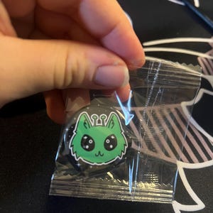 May include: A circular pin badge featuring a stylised green cat head with large black eyes, white highlights, and small wings, set against a black background with white star accents, enclosed in a clear, sealed plastic packaging sleeve.