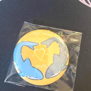 May include: A circular pin-back button featuring a yellow background with a blue shark and a grey shark facing each other to form a heart shape. The design includes small white heart outlines and sparkles, all encased in a clear plastic protective sleeve.