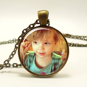 May include: A round pendant necklace featuring a bronze-toned metal frame and a chain. The circular insert displays a photograph of a young child with light brown hair wearing a light blue and green plaid shirt, holding white wired earbuds.