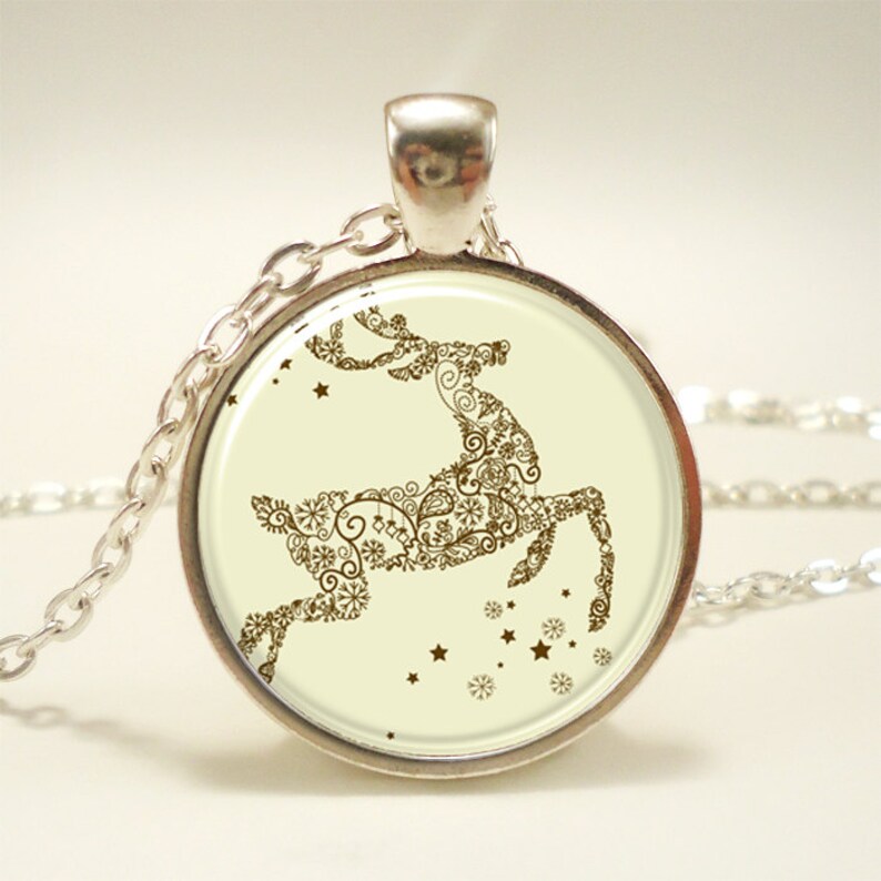 Op de afbeelding: Een zilveren ketting met een ronde hanger met een bruin silhouet van een rendier met ingewikkelde details. Het rendier is omringd door kleine gouden sterren en sneeuwvlokken op een cr&egrave;mekleurige achtergrond.