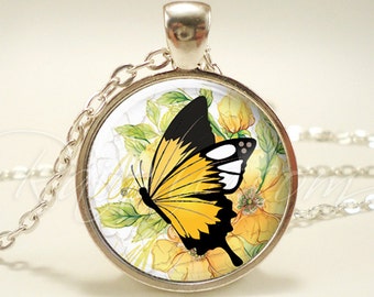 Op de afbeelding: Zilverkleurige hangerketting met een ronde bedel met een vlinderontwerp. De vlinder heeft gele en zwarte vleugels, tegen een achtergrond van gele bloemen en groene bladeren. De ketting is delicaat.