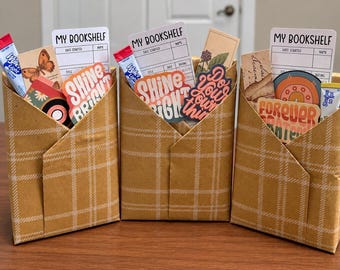 May include: Three tan paper envelopes with a white plaid pattern, each containing a My Bookshelf card, stickers with text like Shine Bright and Do Your Own Thing, and a small drink mix packet. The items are arranged upright in a row.