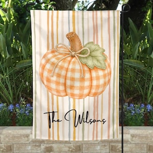 May include: A rectangular garden flag featuring a central orange and white gingham pumpkin with a twine bow and a green leaf. The background consists of vertical stripes in muted tones of orange, yellow, and green. The text "The Wilsons" is printed in black.