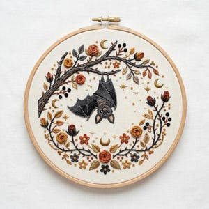 Peut inclure: Un tambour à broder circulaire en bois contenant une pièce de tissu avec un motif cousu main représentant une chauve-souris noire suspendue à une branche. Elle est entourée d'une couronne de roses, feuilles, baies et lunes dorées.
