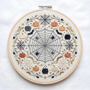 Peut inclure: Un tambour à broder circulaire contenant un panneau en tissu blanc avec un motif détaillé sur le thème d'Halloween. La broderie présente une grande toile d'araignée centrale avec des araignées, entourée de citrouilles orange, de chaudrons noirs et de chauves-souris.