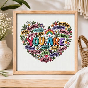 Può includere: Un'opera d'arte a punto croce con cornice quadrata che presenta una composizione a forma di cuore di fiori e foglie colorate che circondano il testo YOU ARE. Sono incluse parole più piccole come beautiful, capable, strong, kind, courageous, resilient, brave e loved.