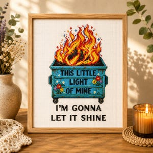 May include: A framed cross-stitch embroidery featuring a blue dumpster engulfed in bright orange and yellow flames. The text reads, "THIS LITTLE LIGHT OF MINE I'M GONNA LET IT SHINE." The design includes small colorful floral accents on the dumpster.