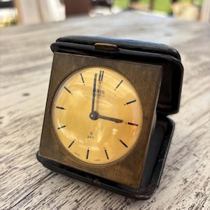 May include: A vintage Oris 8-day travel alarm clock housed in a black leather folding case. The square brass frame surrounds a circular, light yellow analog clock face with black hour markers, black hands, and the text Oris 7 Jewels 8 Day Swiss Made.