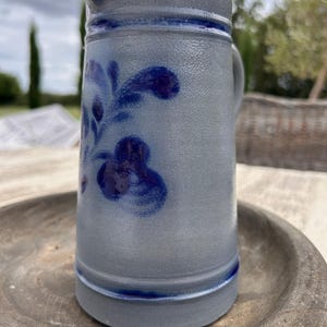 Op de afbeelding: Een grijze stenen kan met een getextureerd oppervlak en een blauw bloemmotief. De kan heeft een opvallende schenktuit, een handvat aan de zijkant en twee horizontale blauwe banden rondom de bovenste en onderste delen van het cilindrische lichaam.