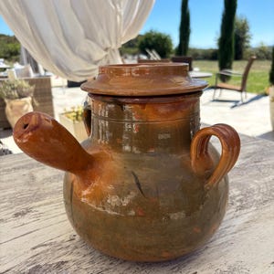 Puede incluir: Una vasija de cerámica vidriada en color marrón con cuerpo redondeado, tapa a juego, un pico vertedor que sobresale por el lado izquierdo y un asa curva en el lado derecho. La superficie presenta tonos tierra moteados con acabado brillante.