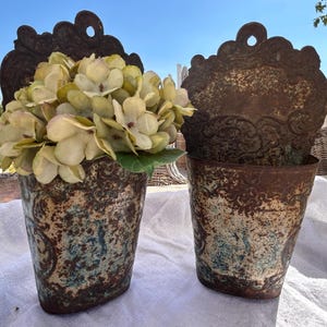 Peut inclure: Deux jardinières murales en métal avec une finition rouillée et patinée, présentant des zones de peinture bleue délavée. Chaque jardinière possède une plaque arrière ornée et festonnée avec un trou circulaire. L'une contient des hortensias vert pâle.