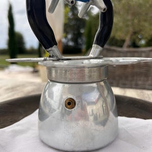 Könnte beinhalten: Ein silberfarbener Espressokocher aus Aluminium mit abgerundetem Boden und einem kleinen messingfarbenen Druckventil. Oben befindet sich ein schwarzer Griff mit einem blau-weißen Etikett, das die Aufschrift Nova Espress MADE IN ITALY trägt.