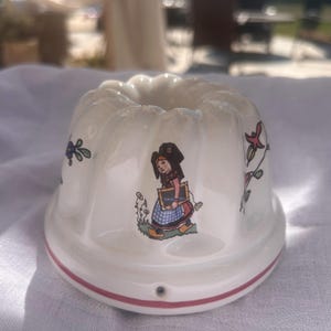 May include: A white ceramic fluted mold featuring a central illustration of a person wearing a traditional cap, a blue and white checkered apron, and wooden clogs, while holding a slate. The mold is decorated with floral motifs and a red line.