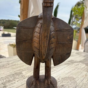 Könnte beinhalten: Eine dunkelbraune, handgeschnitzte Holzskulptur eines Vogels auf einem runden Sockel. Die Figur zeigt stilisierte Flügel mit horizontalen Liniengravuren, einen langen, spitzen Schnabel und einen strukturierten Kopf mit geometrischen Mustern.