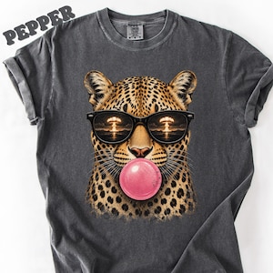 May include: A charcoal gray t-shirt featuring a graphic of a leopard wearing black sunglasses with a reflection of a mushroom cloud. The leopard is blowing a large pink bubblegum bubble. The word PEPPER is printed in the top left corner.
