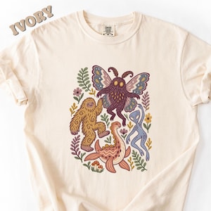 May include: An ivory t-shirt featuring a graphic print of a brown sasquatch, a purple moth, a pink dinosaur, and a blue alien surrounded by colorful flowers and leaves. The shirt has a crew neckline and a Comfort Colors brand label at the neck.