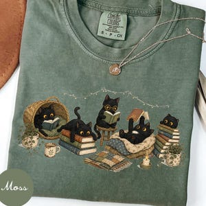Może przedstawiać: Mchowo-zielony t-shirt z grafiką przedstawiającą pięć czarnych kotów czytających książki w przytulnym otoczeniu. Koty siedzą na stosach książek, w wiklinowym koszu oraz na wzorzystym kocu, w towarzystwie małej świeczki.