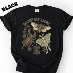 May include: A black t-shirt featuring a graphic print of a brown stag beetle, a tan cicada, and a light green moth. The text "YOU'RE NOT ALONE" is printed in an arch above the insects, and "THERE ARE BUGS" is printed in a straight line below them.