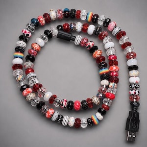 May include: A USB charging cable decorated with a variety of colorful beads in shades of red, black, white, and clear. The beads feature diverse patterns including stripes, glitter, and solid colors, arranged along the length of the black cable.