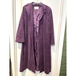 May include: A long, deep purple coat featuring a soft, suede-like texture, a notched collar, and long sleeves. The garment is open at the front, revealing a matching purple satin-finish lining. A white brand label is visible at the inner neckline.