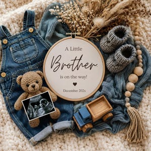 Pode incluir: Um bastidor de bordado redondo de madeira com tecido branco apresentando o texto A Little Brother is on the way! e December 202x. Inclui jardineiras de ganga azul, um pequeno urso de peluche de croché, uma ecografia, sapatinhos de malha cinzentos e um camião de brinquedo de madeira.